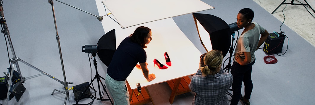 Dos personas en un set fotográfico iluminado capturando un par de zapatos de tacón rojos sobre un fondo blanco