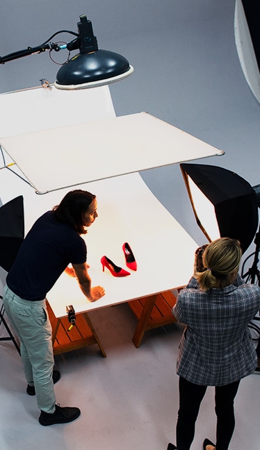 Dos personas en un set fotográfico iluminado capturando un par de zapatos de tacón rojos sobre un fondo blanco