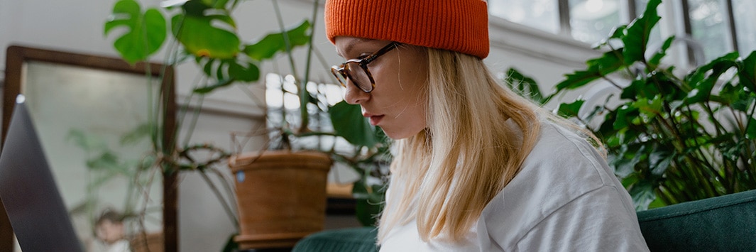 Persona con gafas y gorro escribiendo en un ordenador portátil con plantas y un espejo al fondo