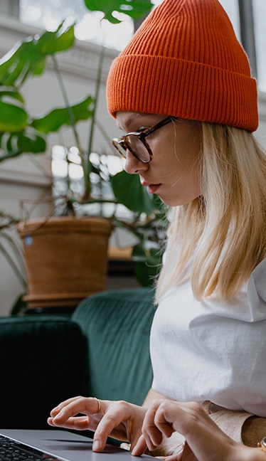 Persona con gafas y gorro escribiendo en un ordenador portátil con plantas al fondo