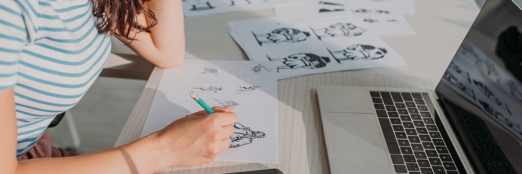 persona ilustrando en papel con un portátil sobre la mesa