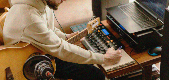 Persona con una guitarra tomando notas en una libreta en un estudio de sonido con una mesa de mezcla y un ordenador